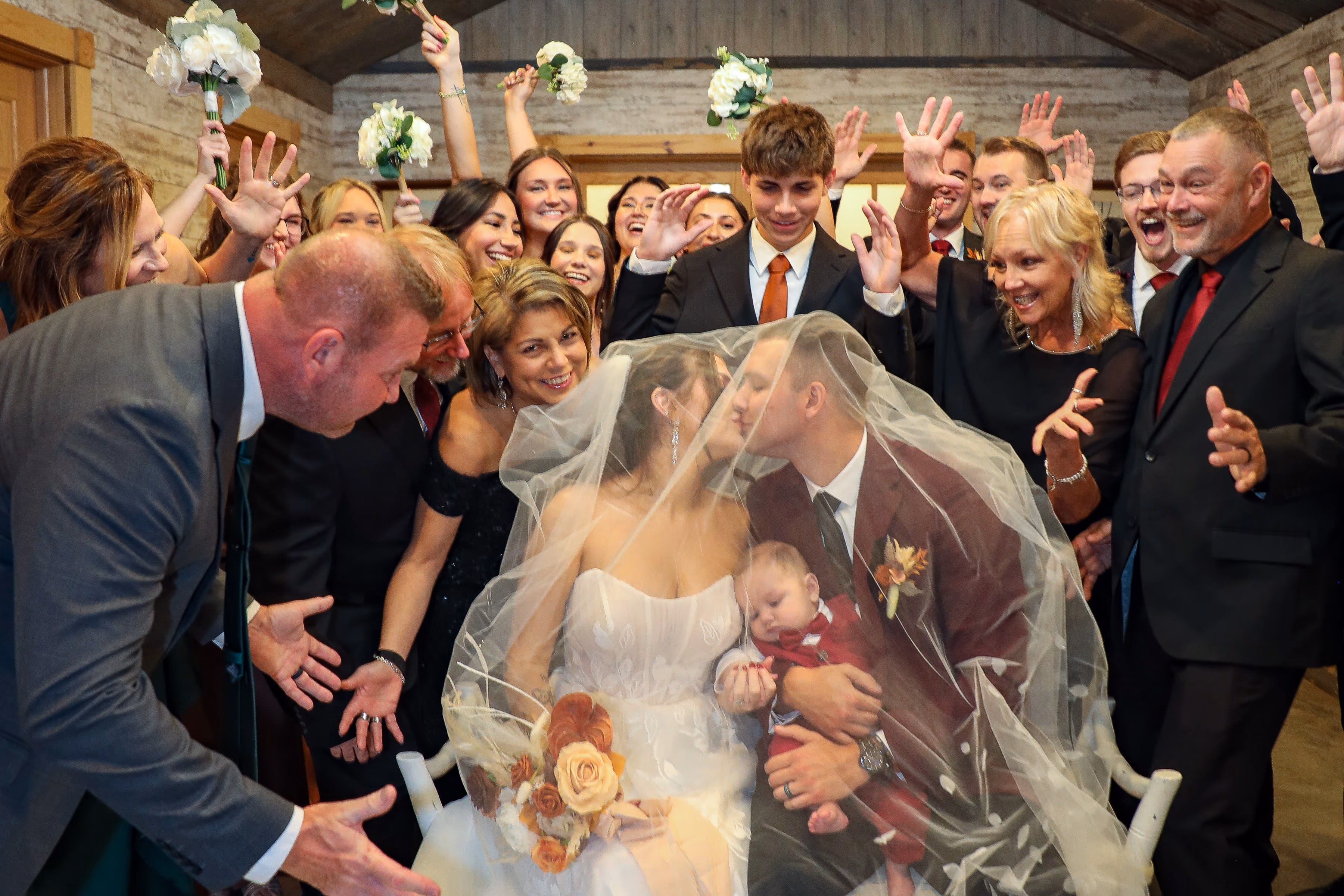 A couple kissing under the veil at their ceremony, guests cheering around them